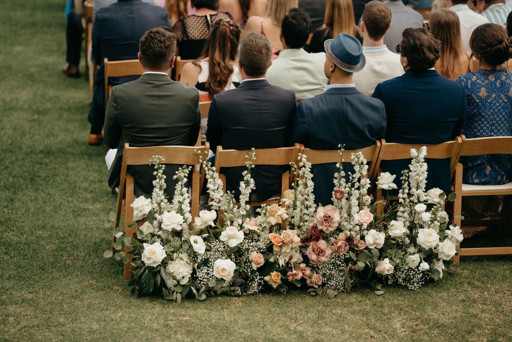 Floral arrangements with white and pink blooms adorn wooden chairs at outdoor wedding ceremony.