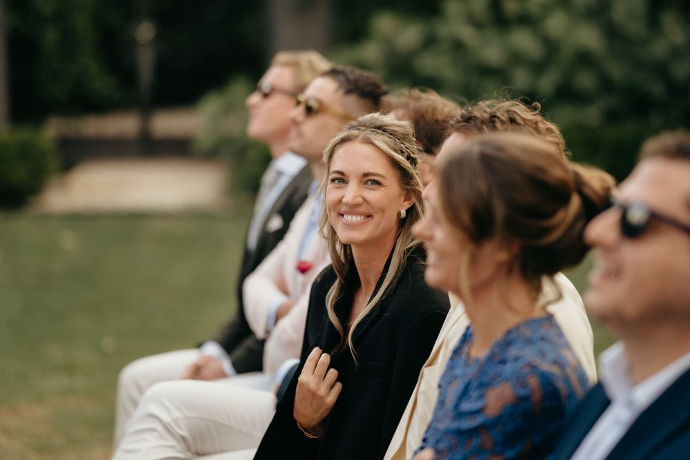 Wedding guests seated in white chairs watch ceremony on a sunny garden lawn.