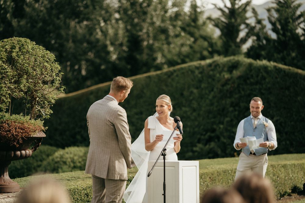 An outdoor wedding ceremony shows speakers at a podium with manicured hedges in the background.