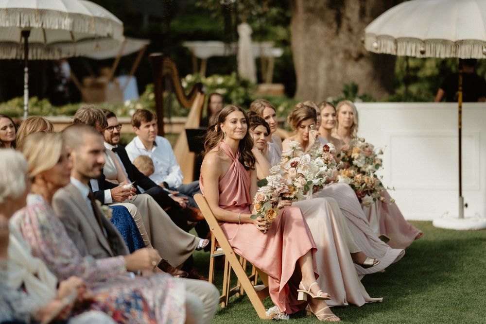 Guests sit in wooden chairs under white umbrellas at an elegant outdoor garden wedding ceremony.