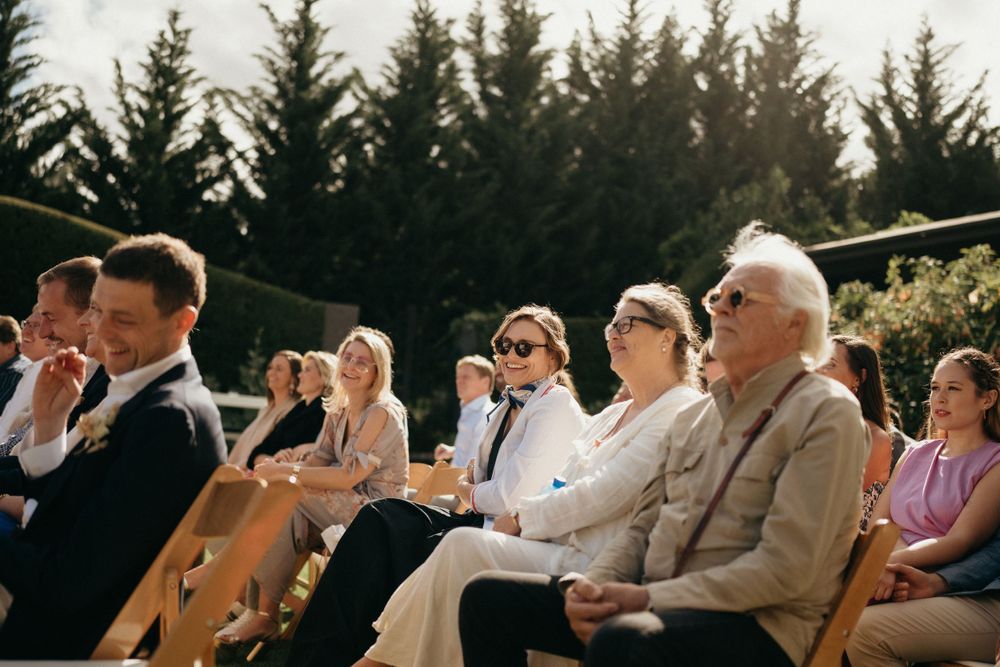 Wedding guests watching ceremony from wooden chairs with evergreen trees in background.