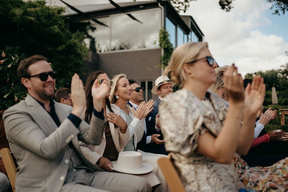 Wedding guests in formal attire applaud during an outdoor celebration.