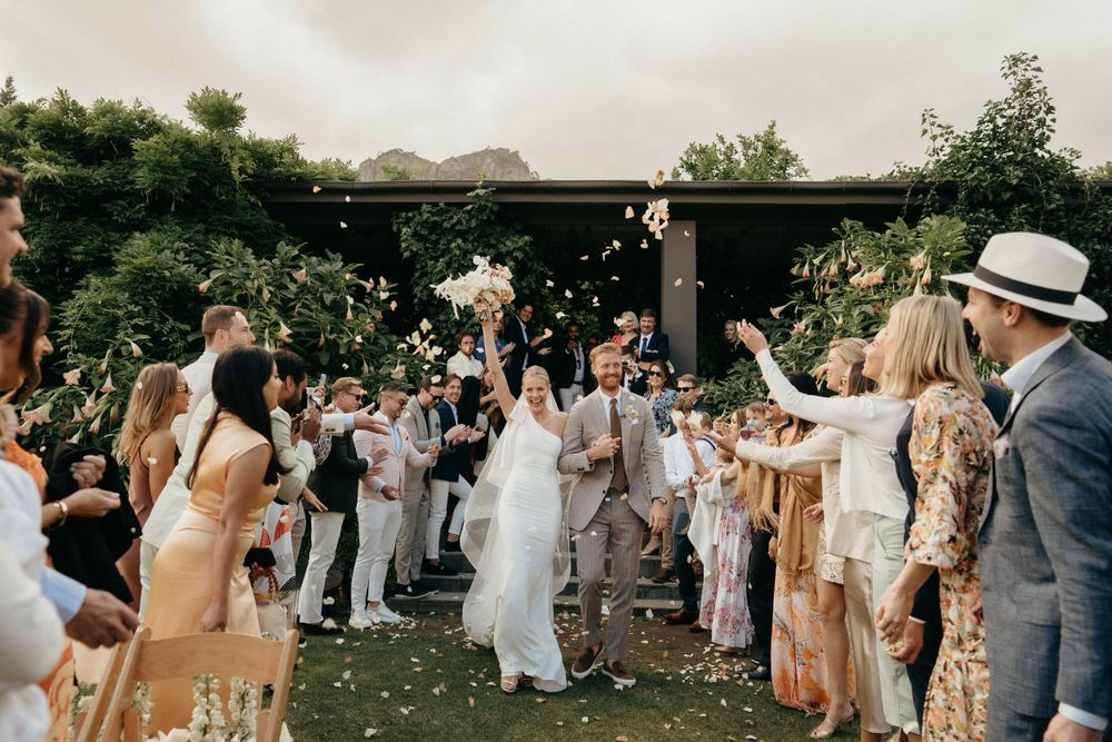 Wedding celebration outdoors with guests in light colored attire tossing flowers and cheering in a garden setting.
