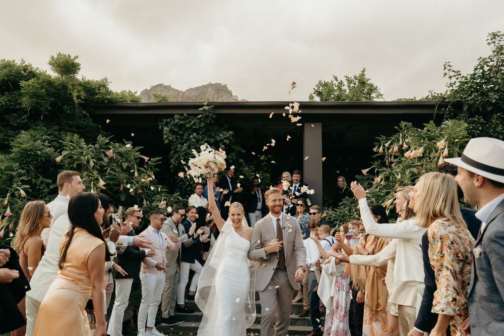 Joyful wedding celebration with guests tossing petals and confetti under an outdoor pergola.