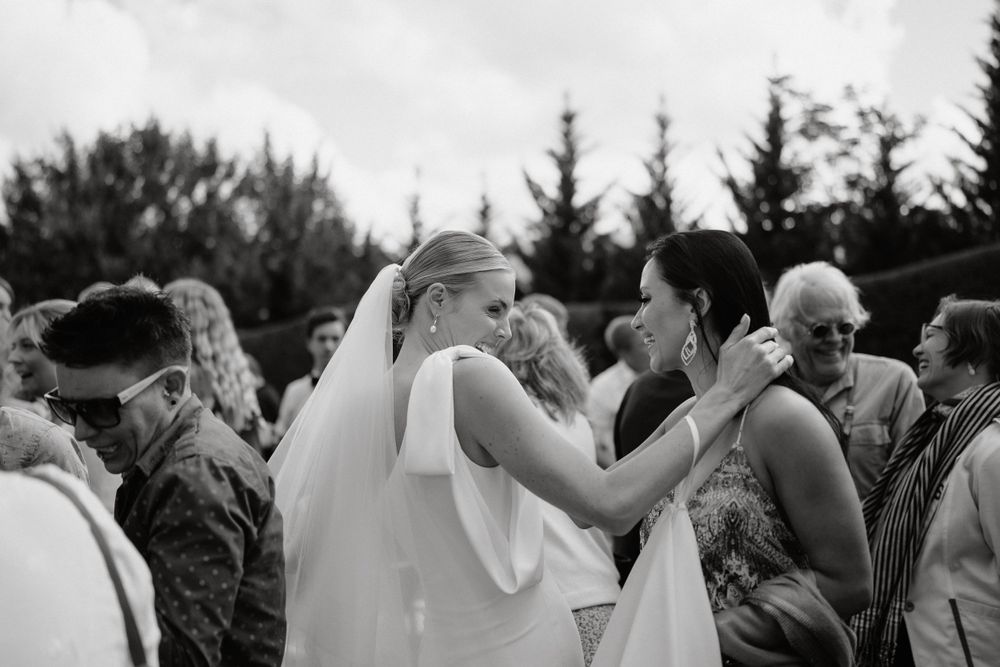 Black and white photo of a bride wearing a veil embracing someone outdoors during a wedding ceremony gathering.