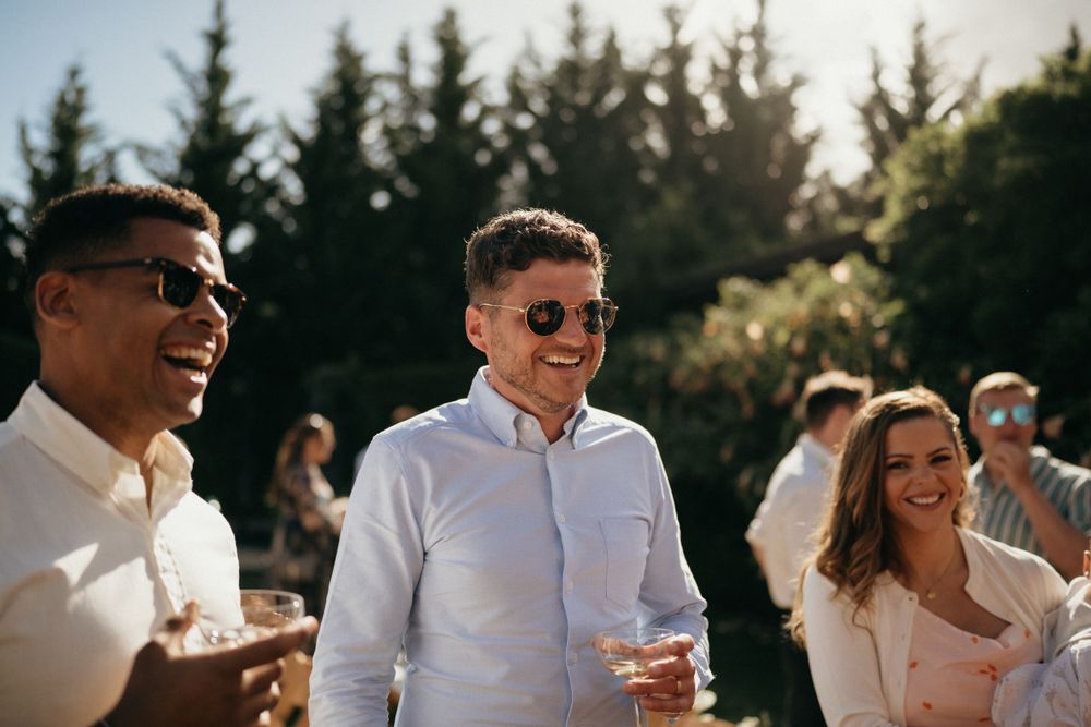 Guests enjoying drinks and conversation at an outdoor wedding reception with evergreen trees in background.