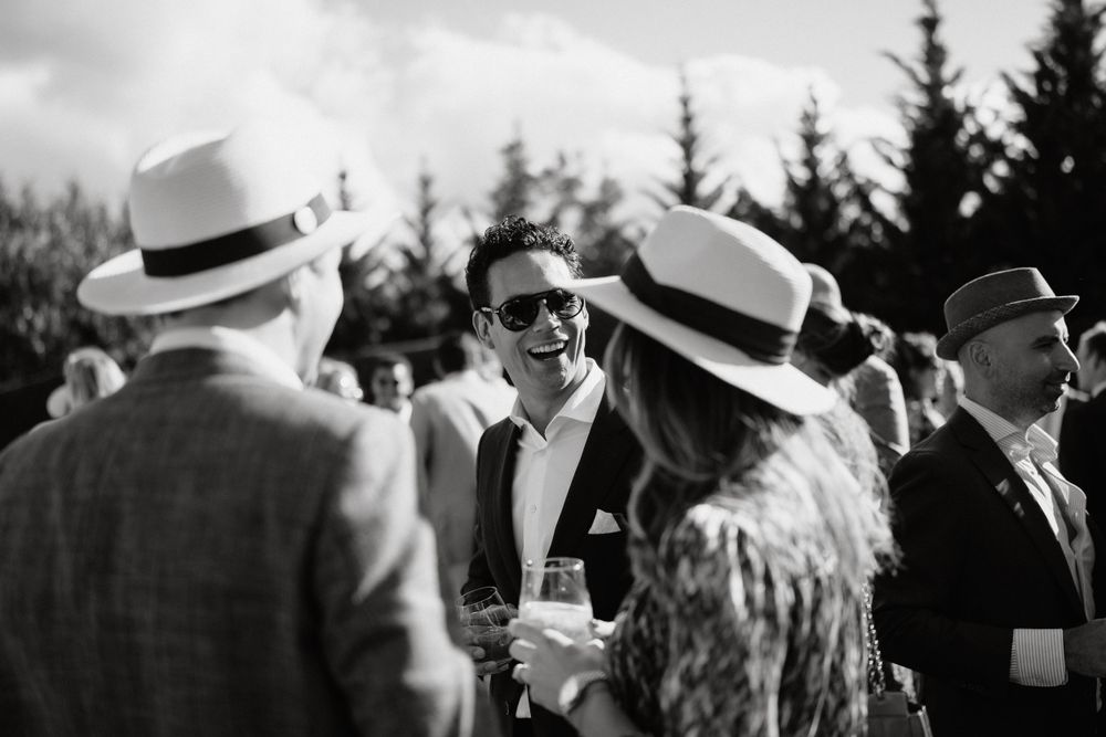 Wedding guests mingle during an outdoor reception wearing stylish hats and formal attire.