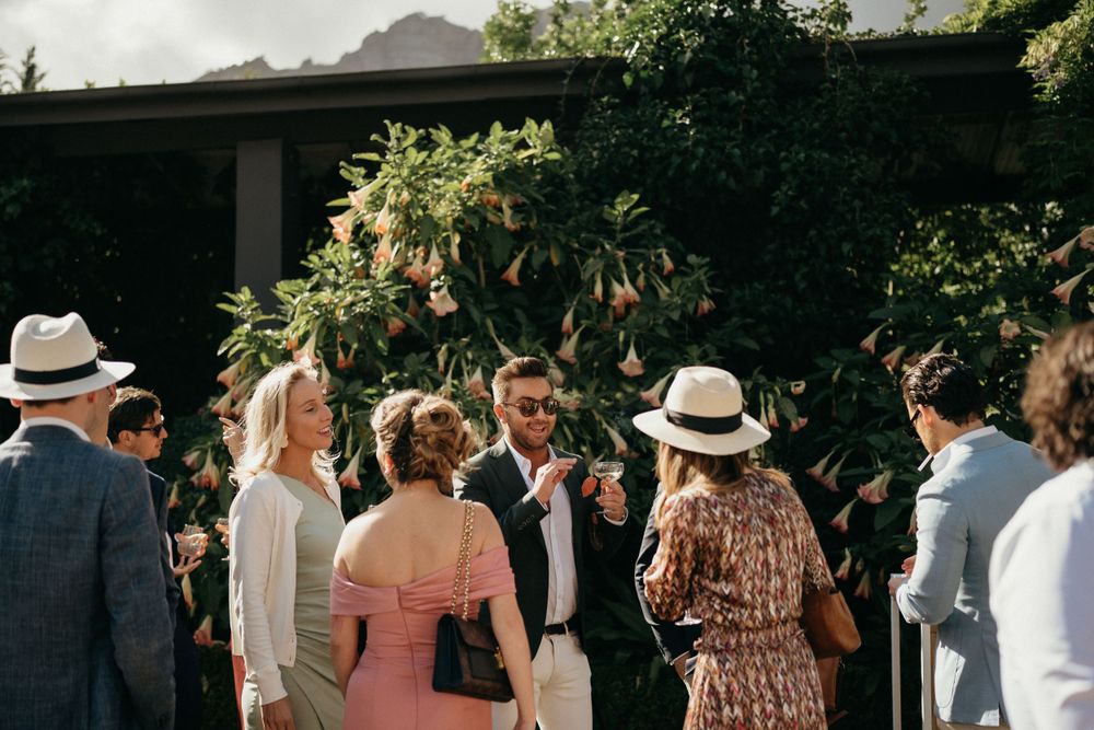 Wedding guests gather outdoors in summer attire around floral decor and greenery during a sunny afternoon celebration.