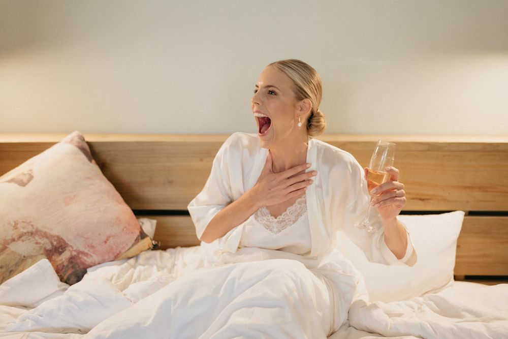 Woman in white lounging in bed with champagne glass expressing joyful surprise.