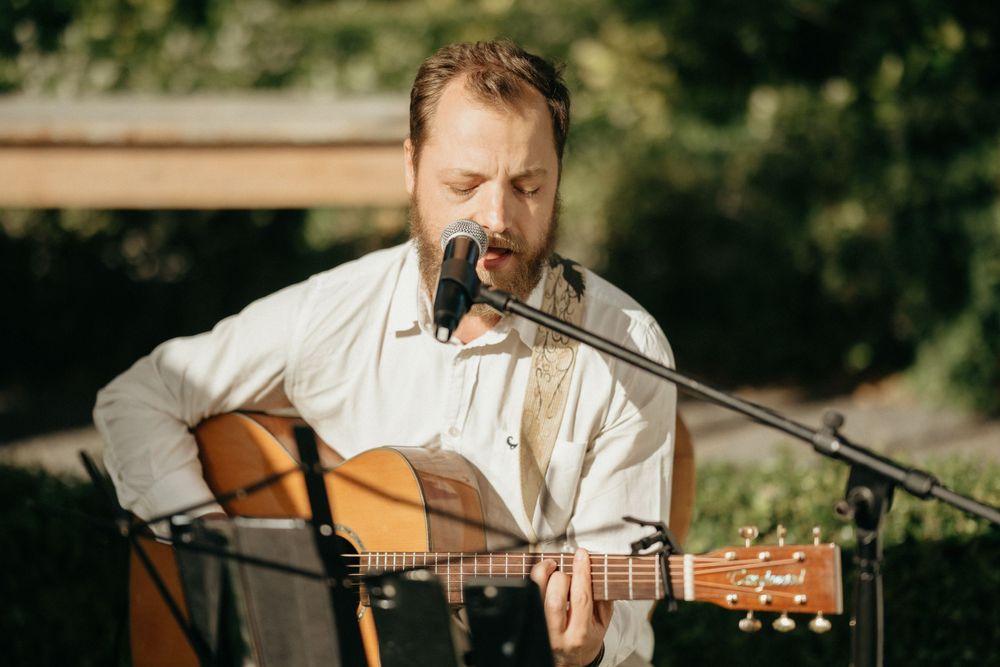 A musician performs outdoors with an acoustic guitar and microphone in a natural garden setting.
