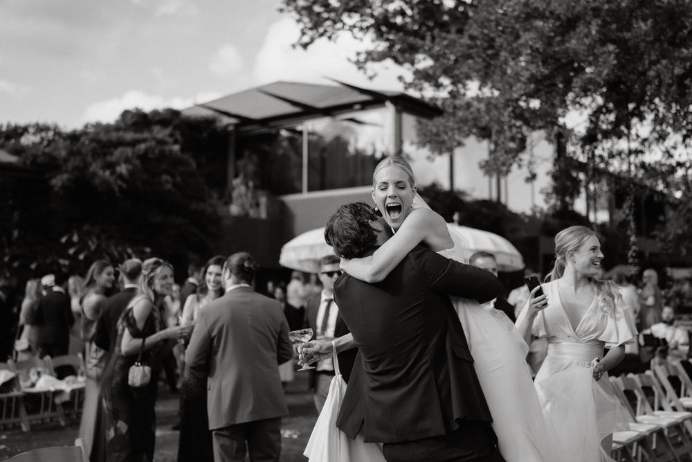 A monochrome photo shows people celebrating at an outdoor wedding reception with guests dancing under trees.