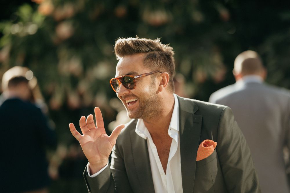 A stylish wedding guest in a grey suit and sunglasses enjoying an outdoor celebration.
