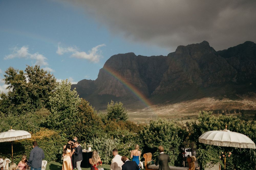 Outdoor wedding celebration with dramatic mountain backdrop and rainbow in the distance.