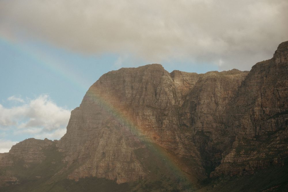 A dramatic rainbow appears against rugged mountain cliffs under cloudy skies.