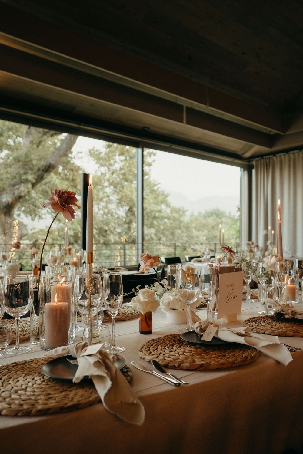 Elegantly set wedding reception table with rustic placemats and floral centerpieces by large windows.