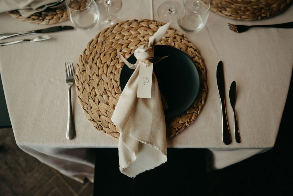 Table setting with woven placemat, black plate, and cream napkin on white tablecloth.