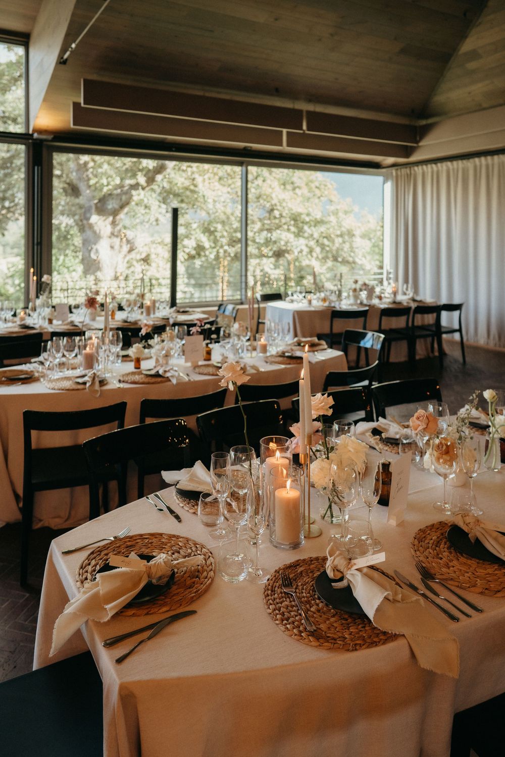 Elegant wedding reception table setting with white linens, woven chargers, and candlelight in a windowed dining room.