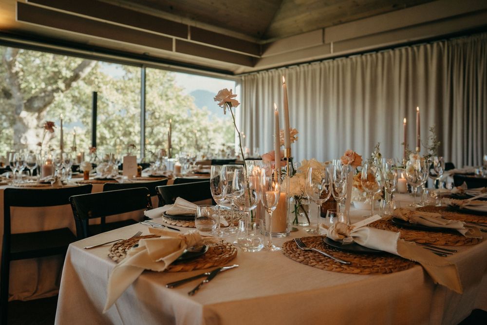 A romantic reception table setup with white linens, glassware and warm candlelight in a windowed venue.