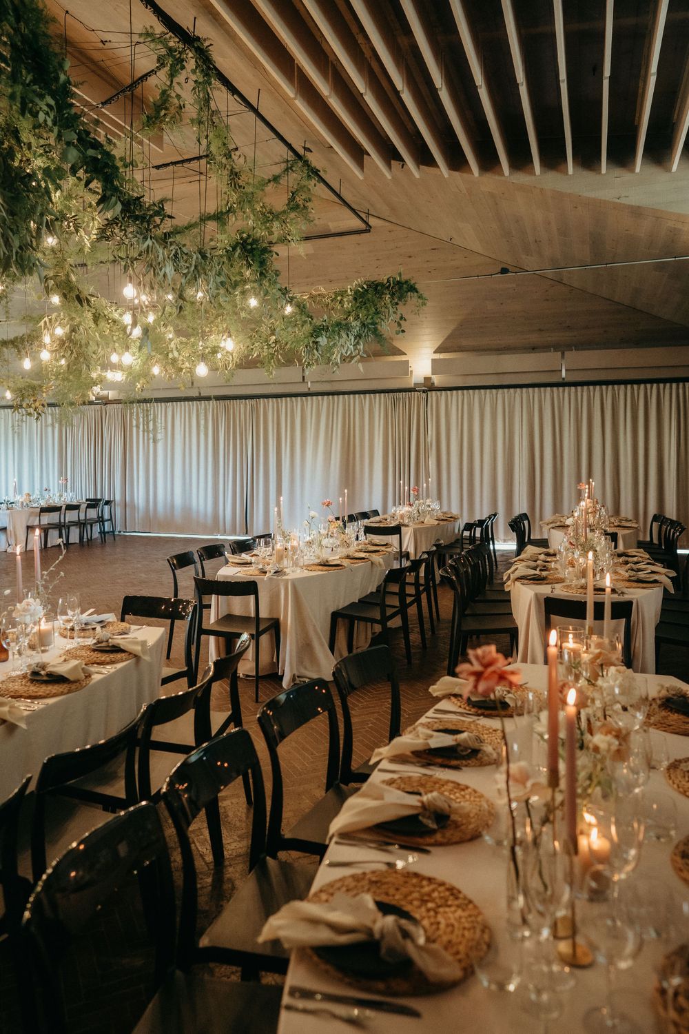 Elegant wedding reception setup with hanging greenery, string lights, and white tablecloths in an industrial-style venue.