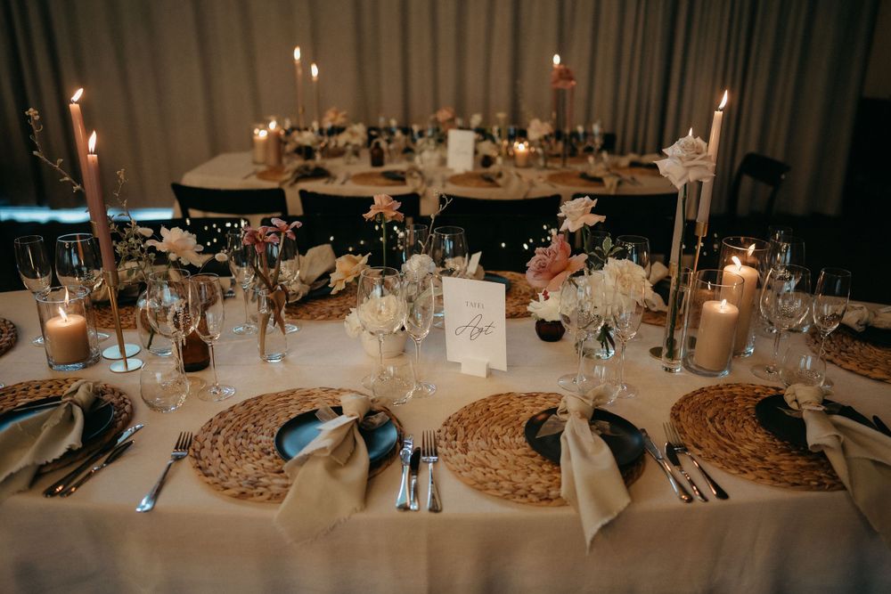 Elegant wedding reception table setting with woven placemats, taper candles, and white tablecloths in moody lighting.