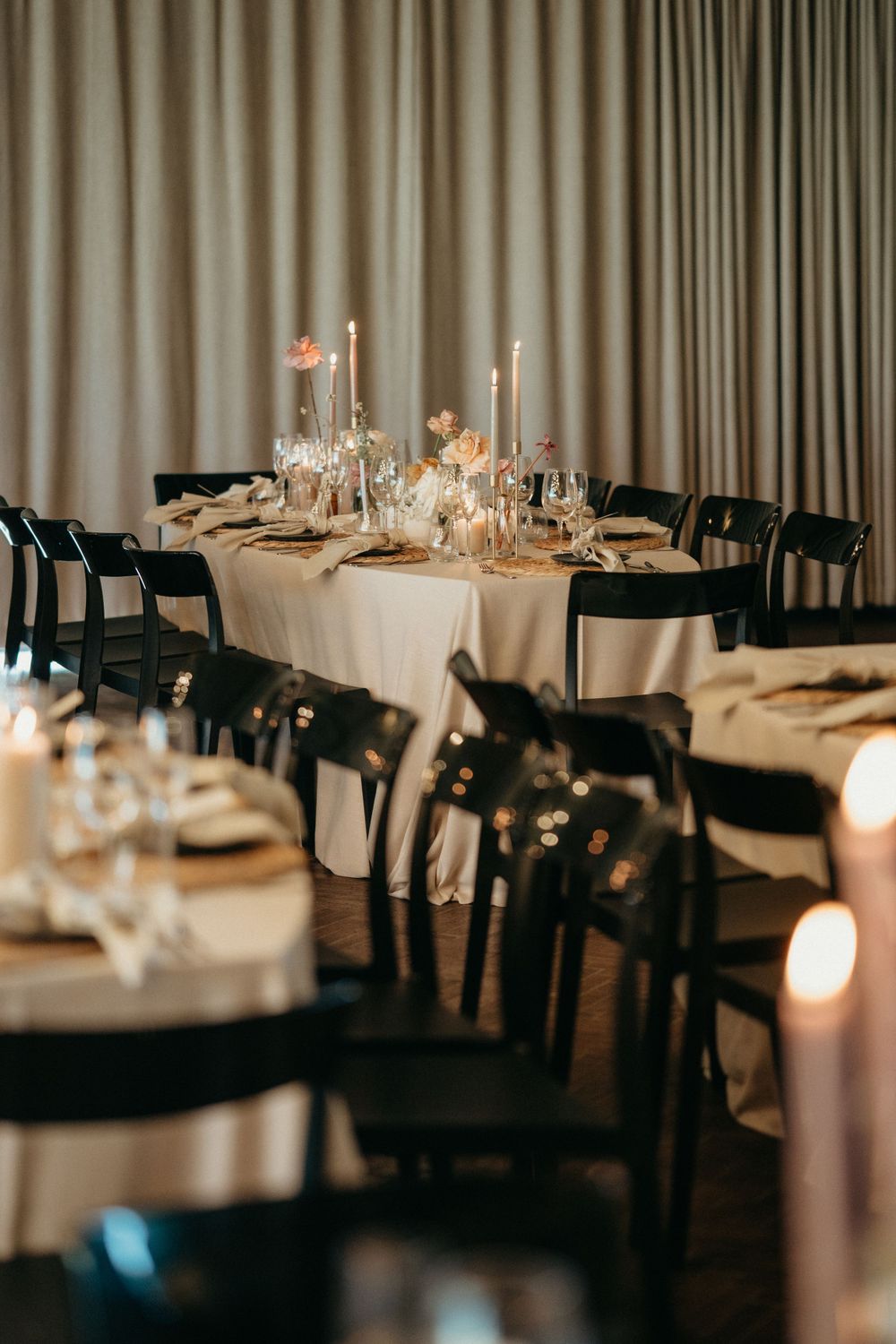 Elegant wedding reception table setting with glowing candles, white linens and black chairs in dimly lit room.