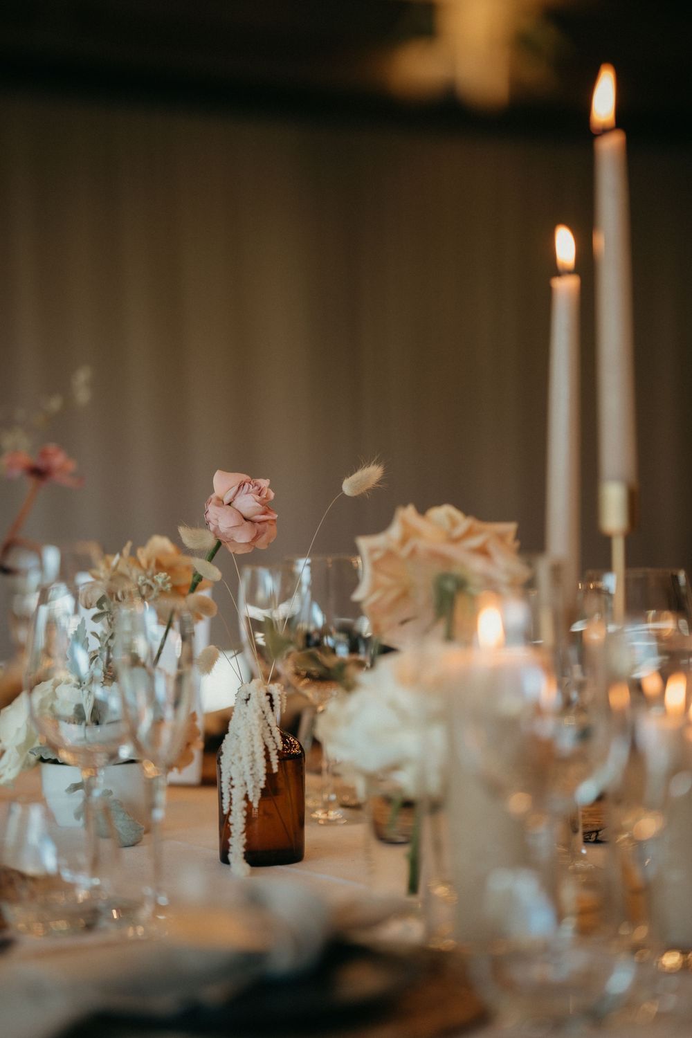 Elegant wedding reception table setting with lit white candles, soft pink flowers and amber glass bottles in romantic lighting.