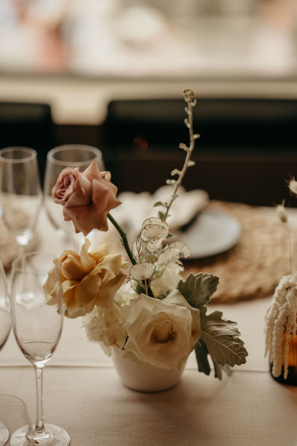 Elegant floral centerpiece with blush roses and white flowers in a small vase on a formal dining table with champagne glasses.