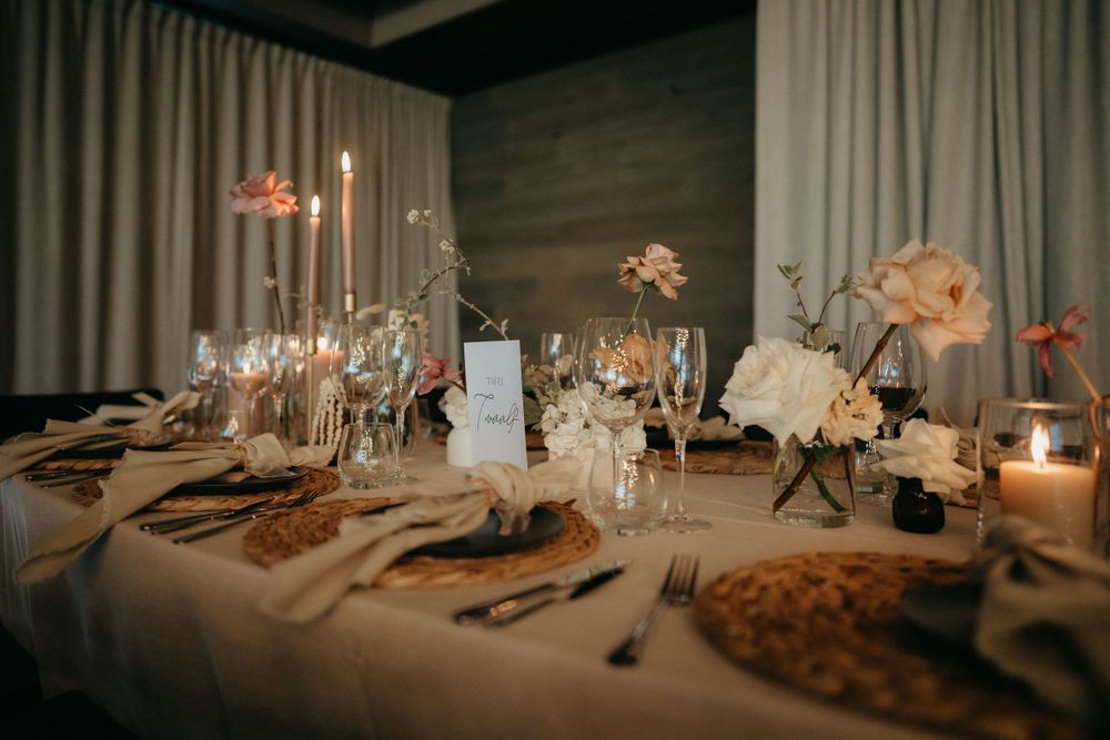 An elegant wedding reception table setting with candles, white floral centerpieces, and place settings on a beige tablecloth.