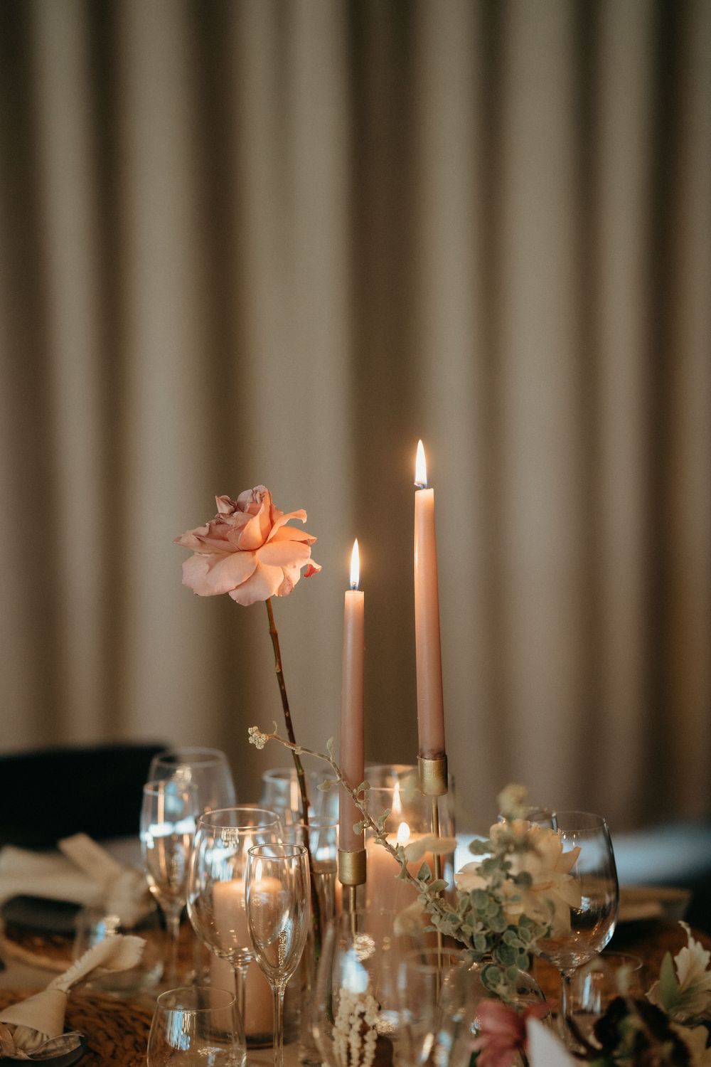 Romantic table centerpiece with tall taper candles and pink rose in vase against soft lighting.