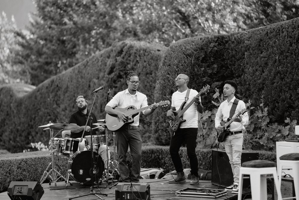 Black and white photo of a live band performing outdoors with instruments against a hedge backdrop.