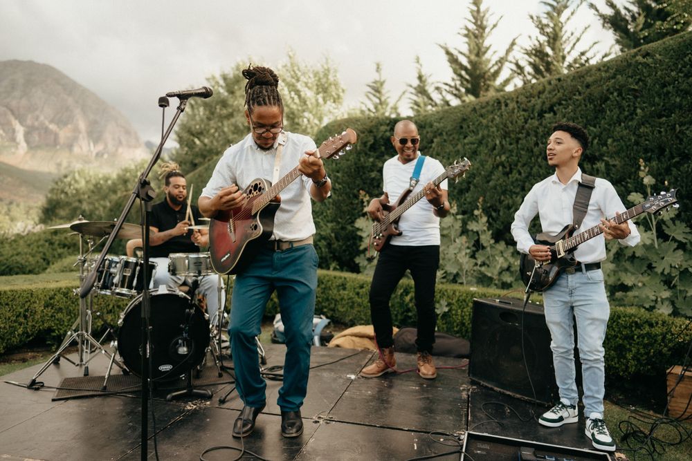 Musicians perform outdoors with guitars and drums against a mountain backdrop during a live event.