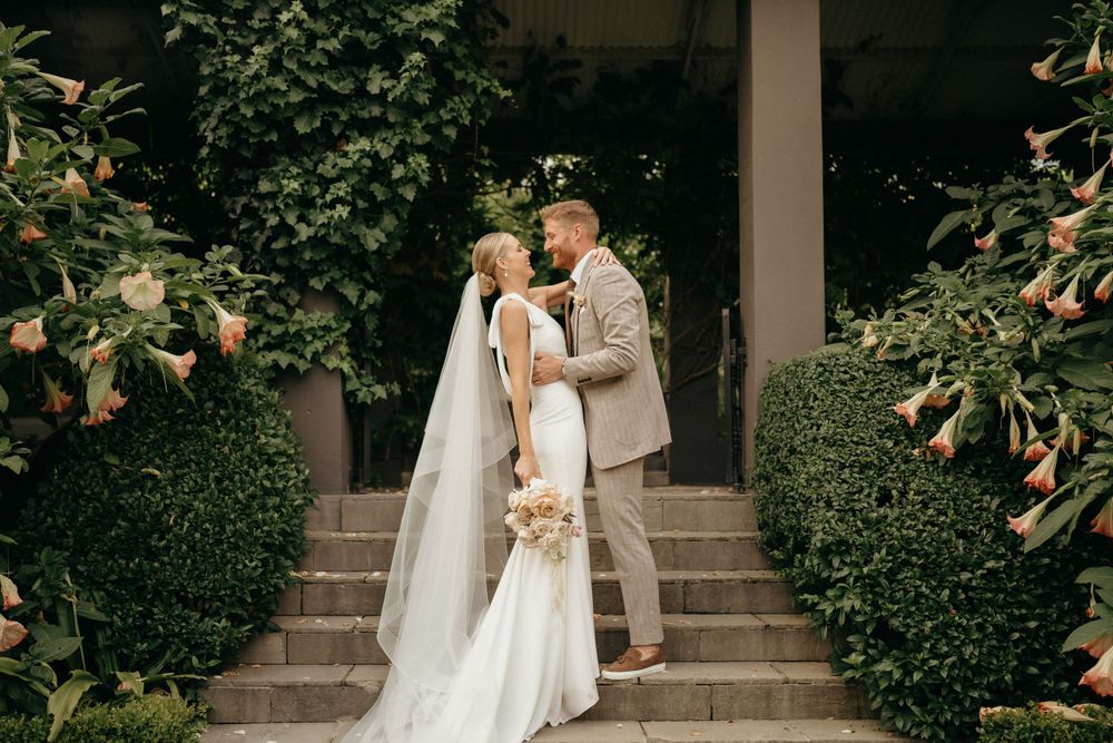 A wedding couple poses elegantly on garden steps framed by blooming shrubs.
