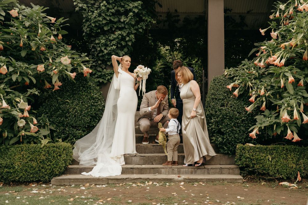 A wedding party poses on garden steps surrounded by lush greenery and floral arrangements.