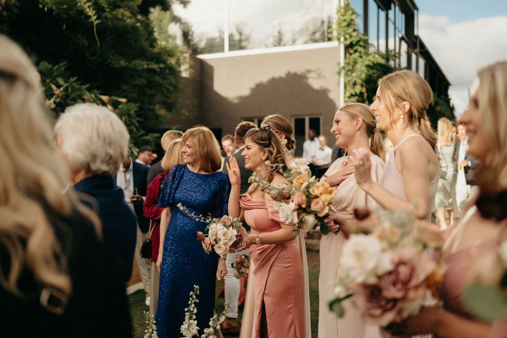 Wedding guests in colorful dresses gather outdoors holding floral bouquets on a sunny day.
