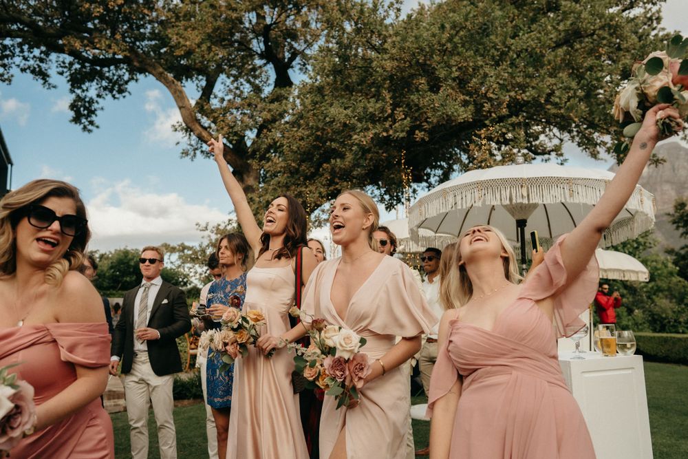 Wedding party in blush pink dresses celebrates joyfully on a sunny outdoor lawn.