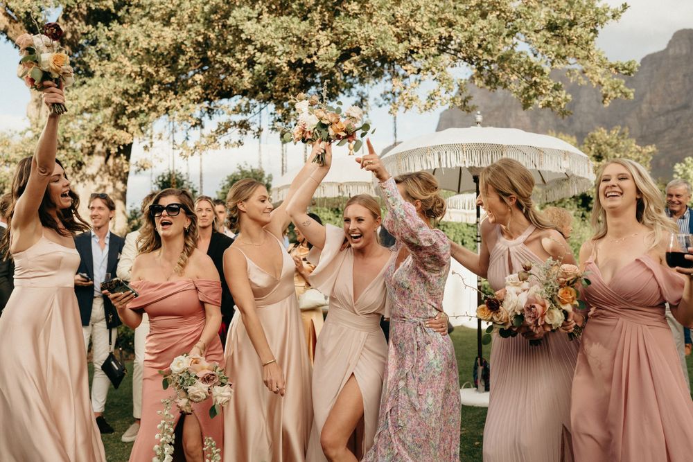 Wedding party wearing blush pink dresses celebrates outdoors under trees with parasols.