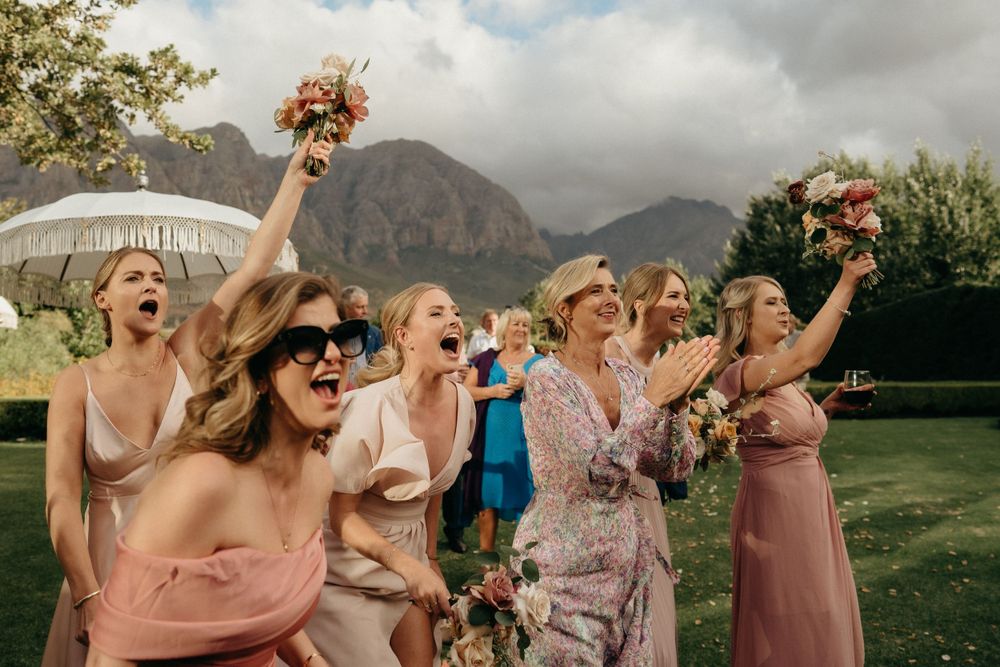 Wedding party celebration on lawn with mountains in background, holding bouquets and parasols.