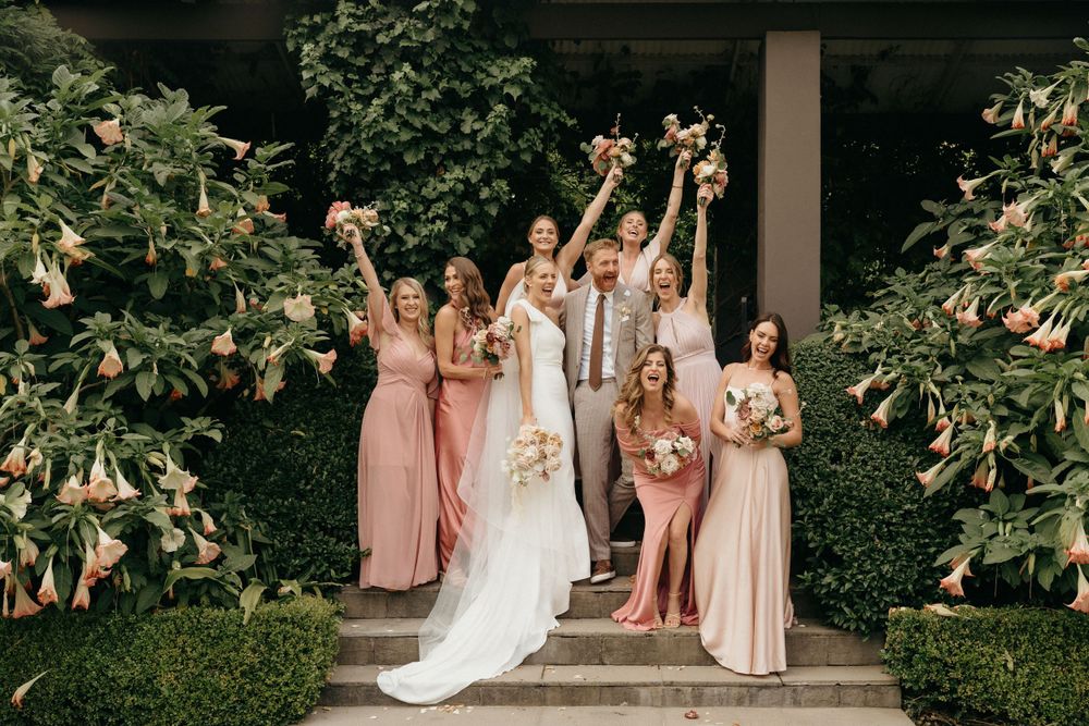 Wedding party in pink dresses posing on garden steps with floral decor.