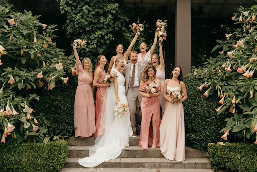 Wedding party in pink and blush dresses posing with bouquets on stone steps in garden setting.