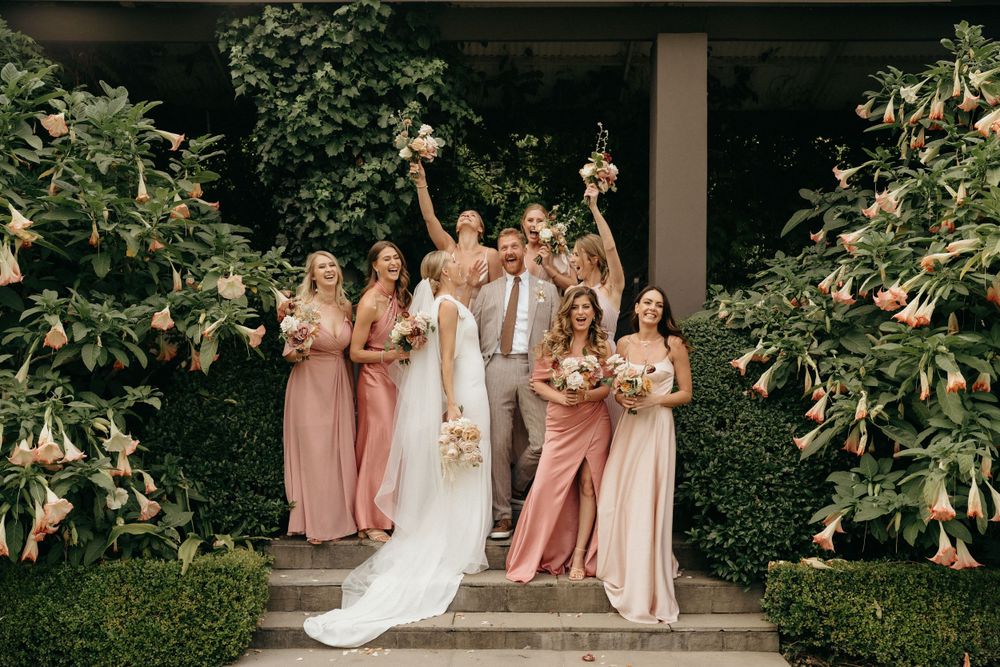 Wedding party in pink and neutral dresses pose together on outdoor garden steps surrounded by greenery.
