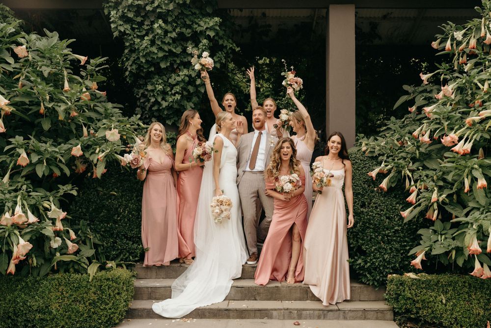 A bridal party in blush and dusty rose dresses poses on stone steps surrounded by lush greenery.