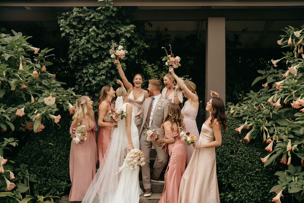 Wedding party celebrates joyfully in a garden setting with bridesmaids in pink dresses.