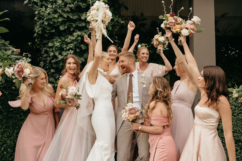 Wedding party celebrating with floral bouquets in pink and blush dresses against a garden backdrop.