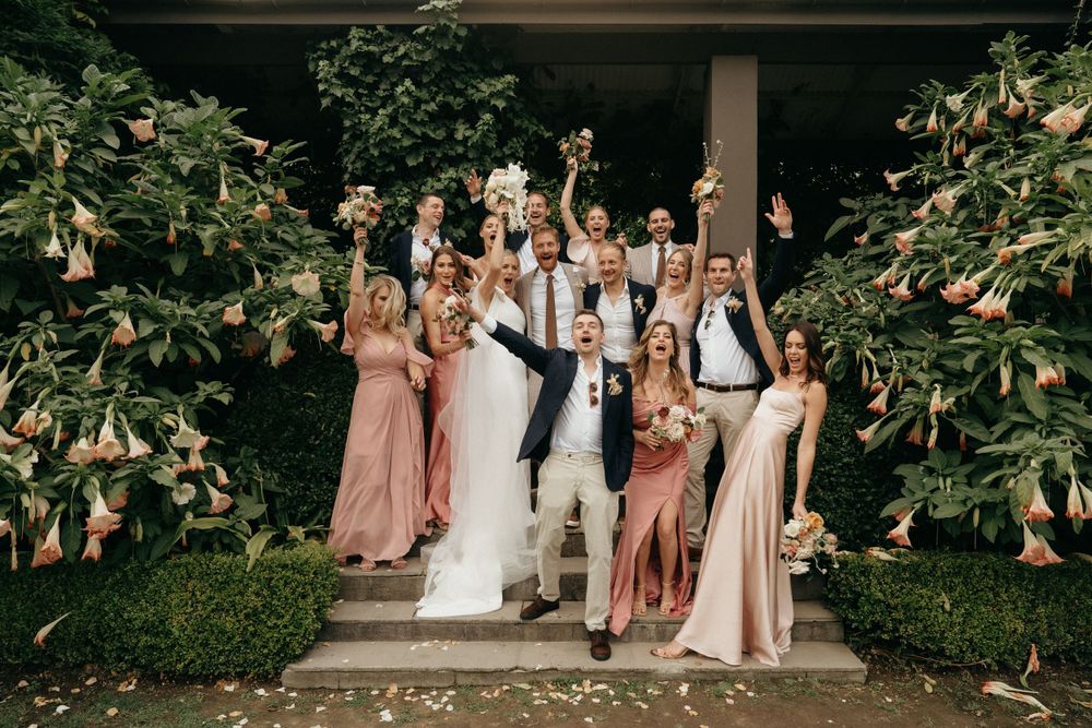 Celebratory group shot of wedding party on outdoor steps with raised bouquets.