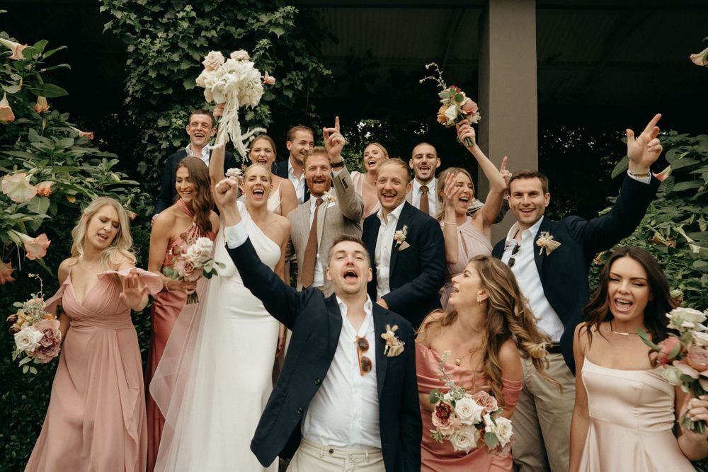 Wedding party celebrates joyfully with raised arms while wearing blush pink dresses and dark suits in an outdoor garden setting.