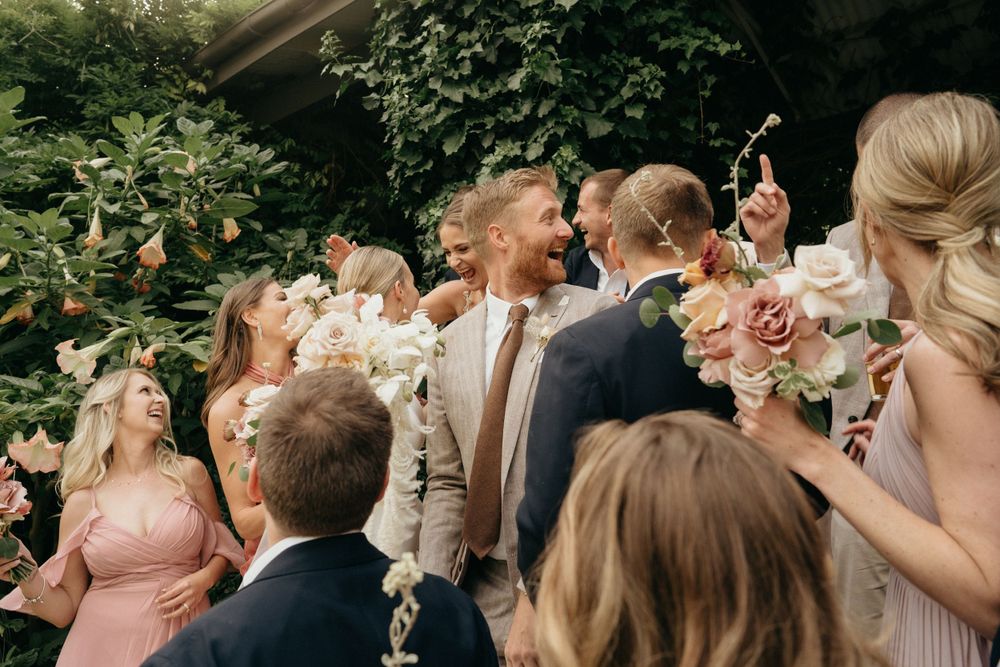 Wedding group shares a candid moment of laughter surrounded by greenery while holding floral bouquets.