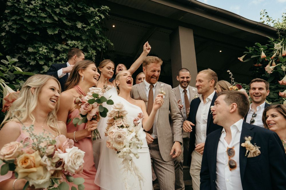 Celebratory moment captured at outdoor wedding with bridal party in pink dresses cheering with bouquets.