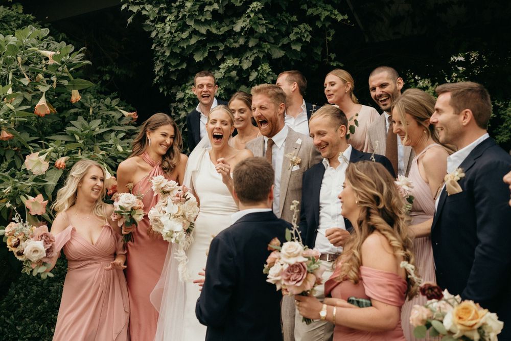 Wedding party gathered outdoors wearing blush pink bridesmaid dresses and light colored suits with floral bouquets.