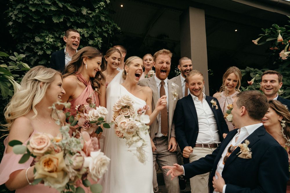 Joyful wedding party gathered outside in formal attire with pink and blush floral bouquets.