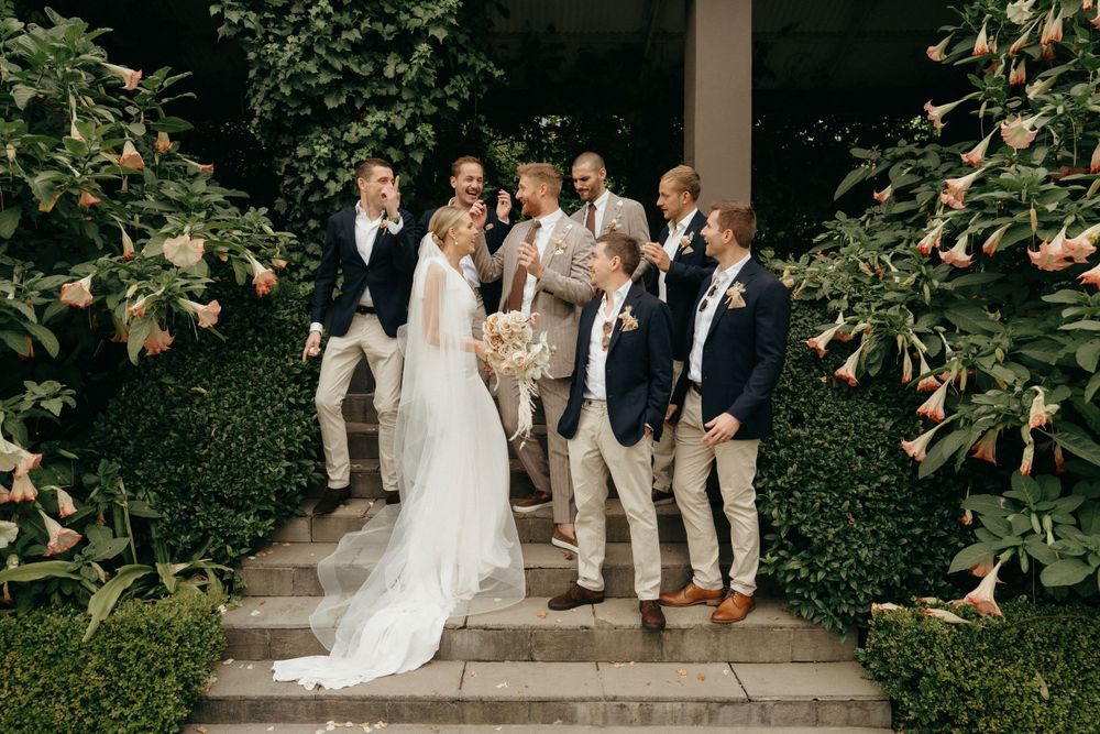 Wedding party in casual formal attire poses on garden steps with lush greenery backdrop.